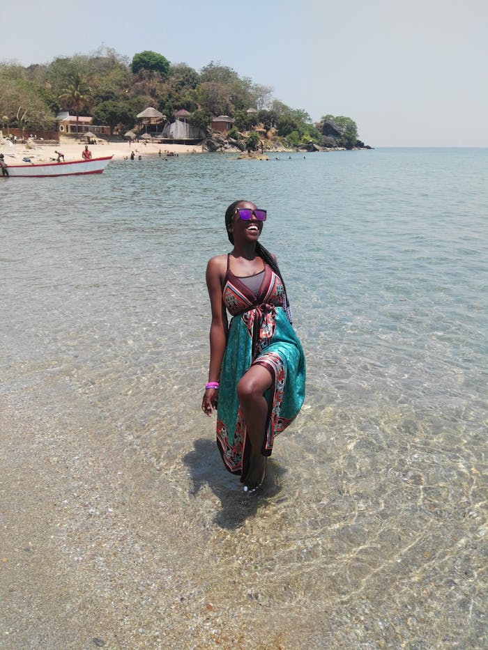 Smiling woman in a colorful dress enjoying a sunny day at the beach, standing in clear water.