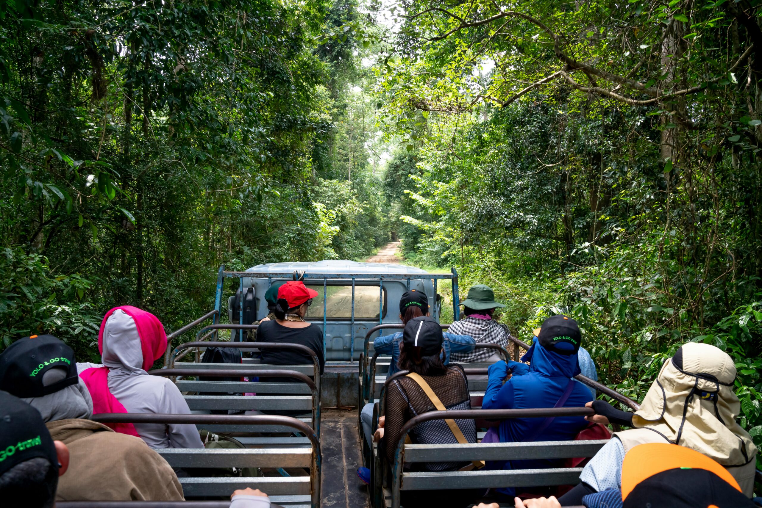 Anonymous travelers in headwear sitting on seats of truck driving through tropical forest